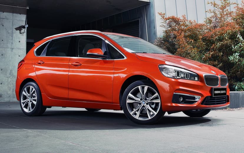 Metallic orange BMW 2 Series parked in front of a minimalist concrete building with autumn foliage background.