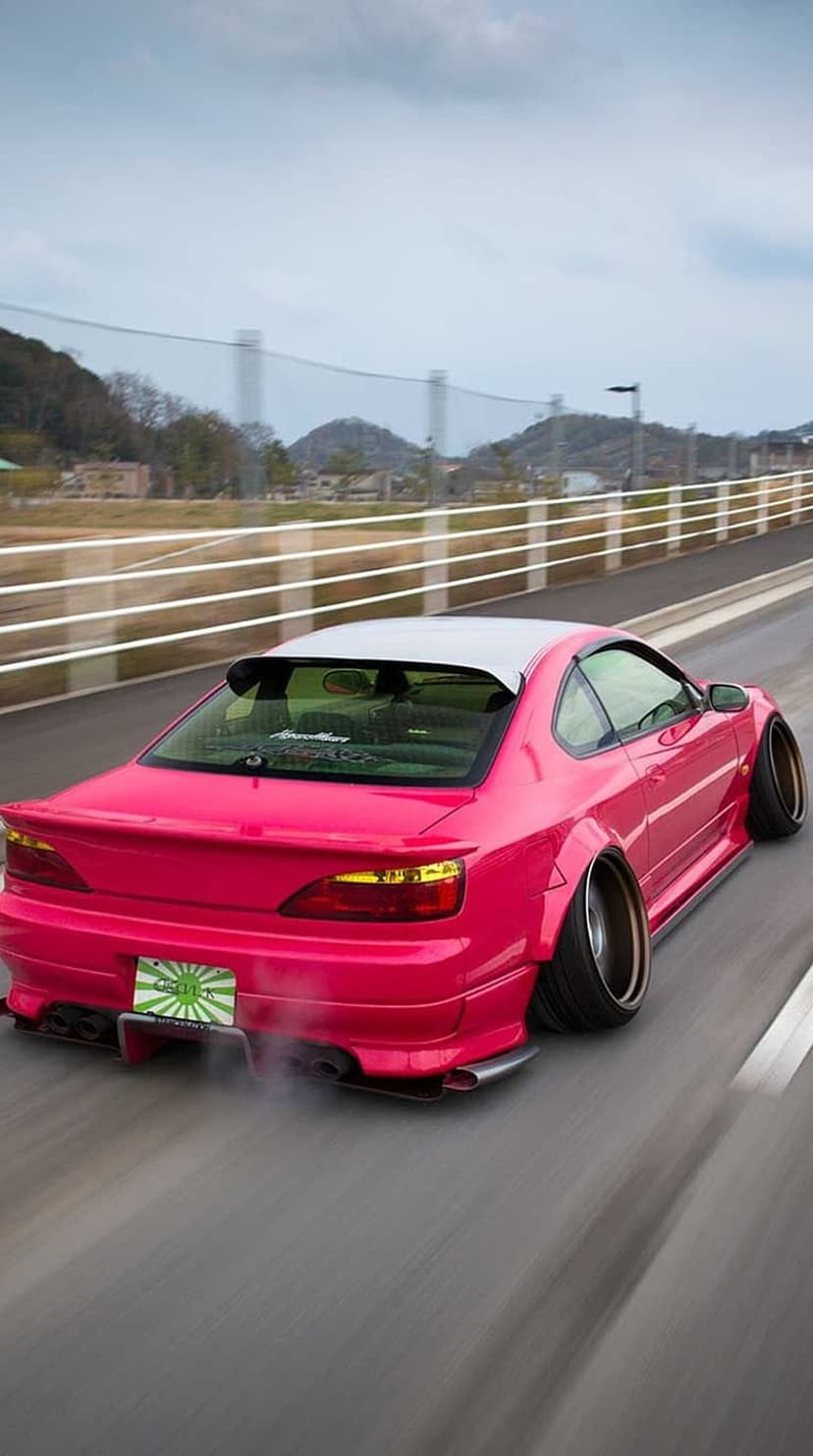 Pink Nissan Silvia S15 drifting with white tire smoke on a paved mountain road against a cloudy horizon.