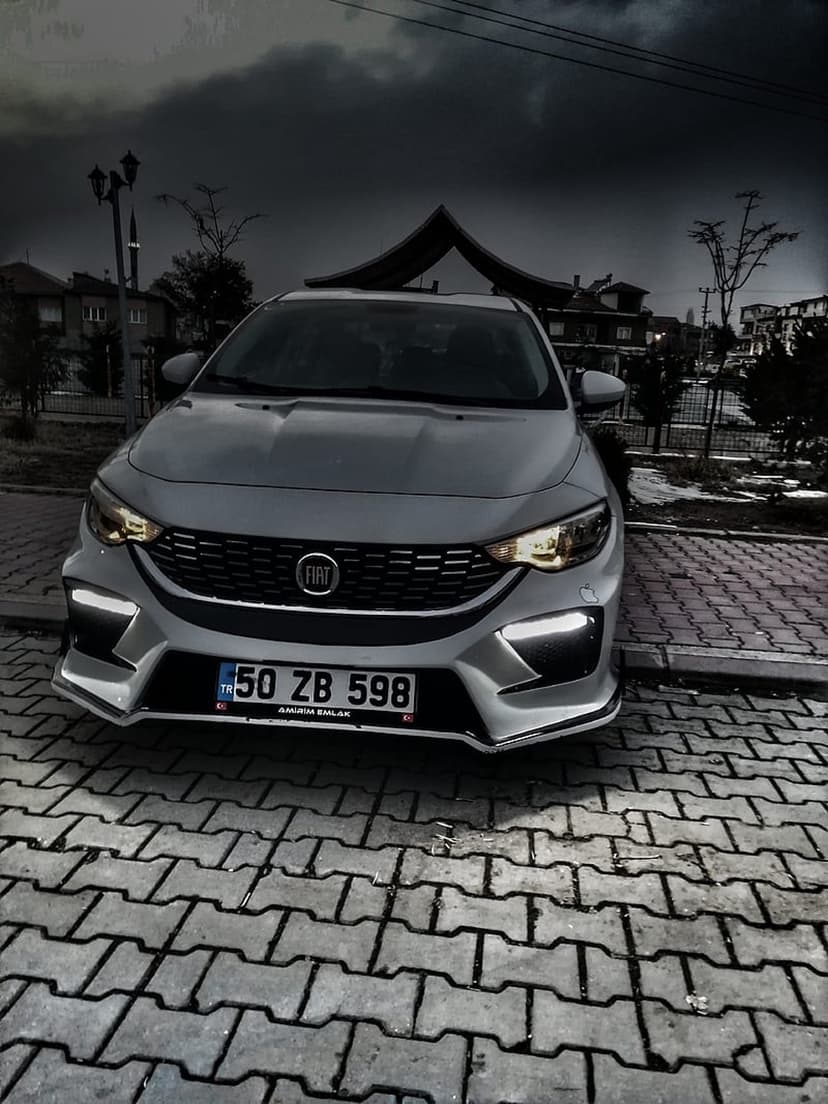 Silver Fiat sedan with glowing headlights parked on a brick pavement under dark, dramatic stormy clouds at dusk.