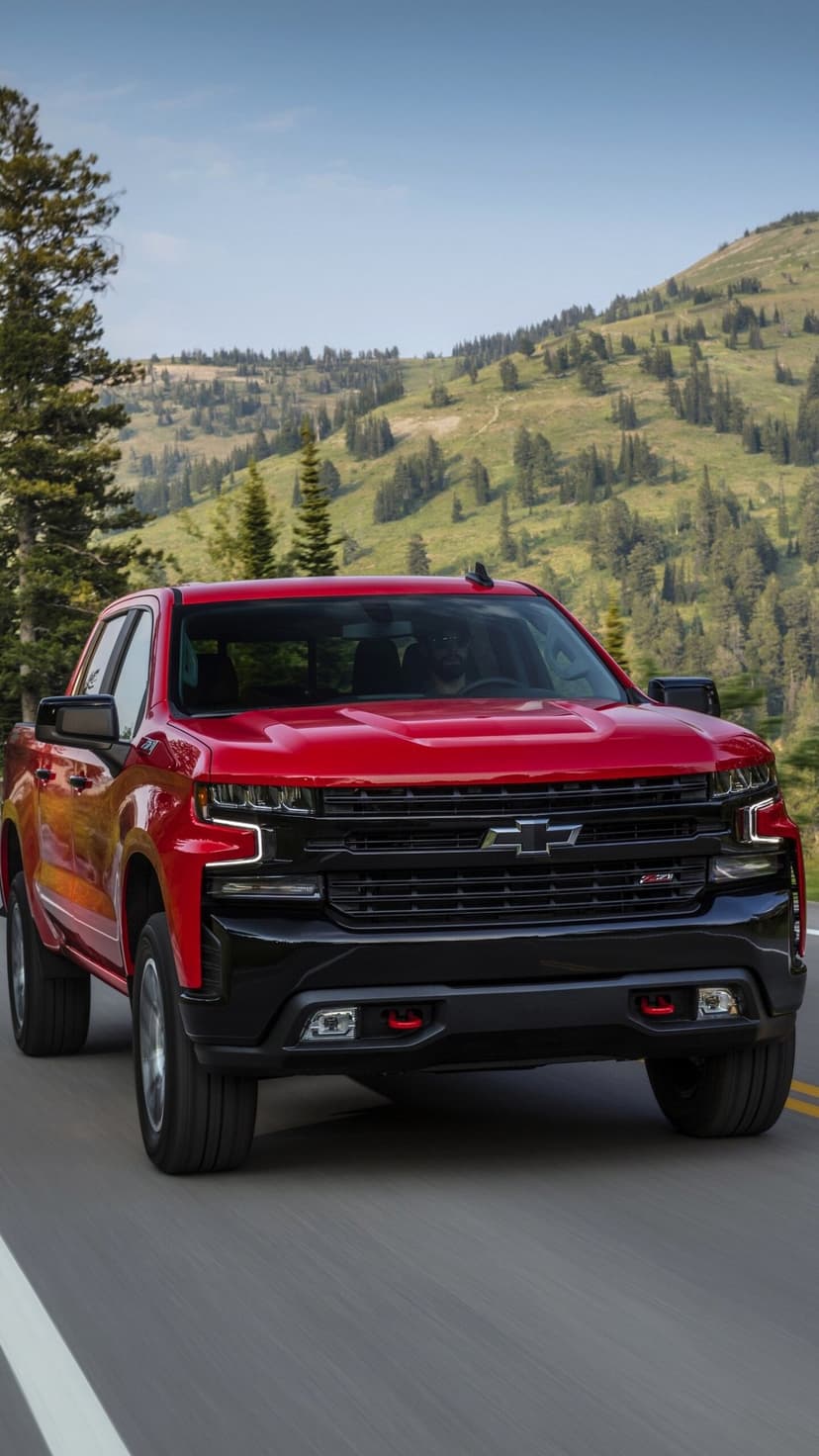 Red Chevrolet Silverado truck driving on a mountain road with green forests and hills in the background.