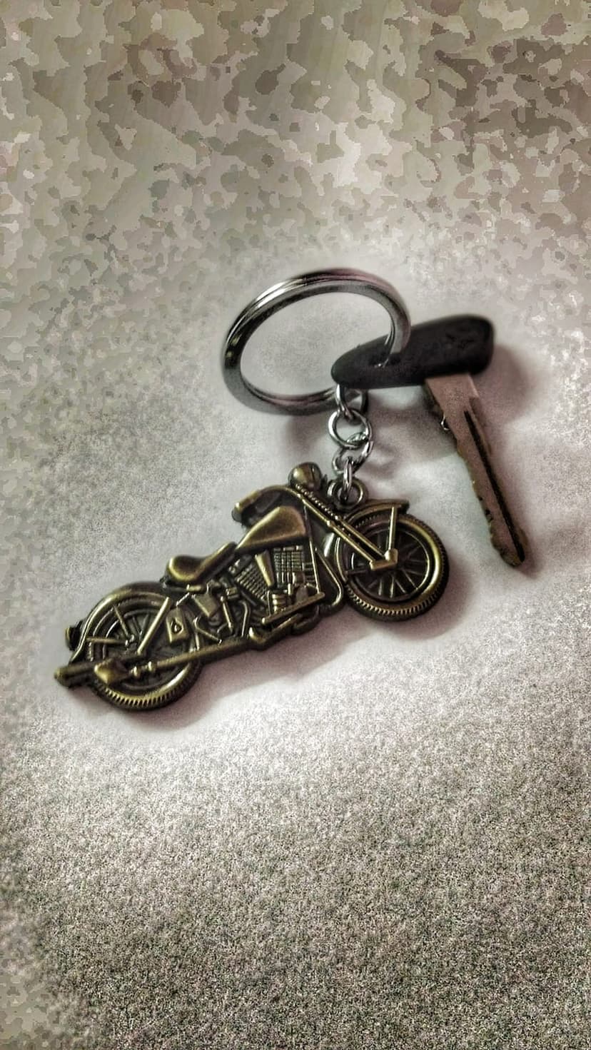 Metal motorcycle keychain and a car key resting on a light-colored speckled surface with soft lighting.