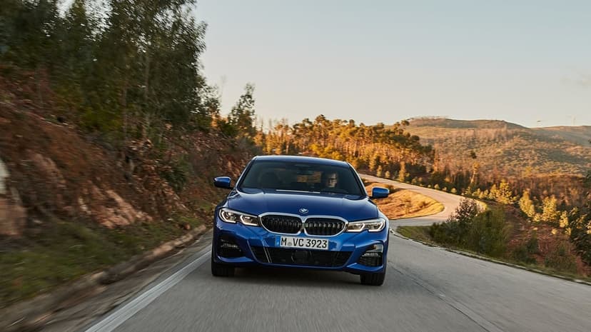 Front view of a blue BMW sedan driving along a winding mountain road during golden hour with sun-drenched hills.