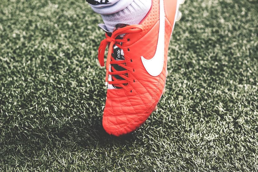 Orange Nike soccer cleat with white laces sits on green artificial turf blades at a low-angle macro perspective.