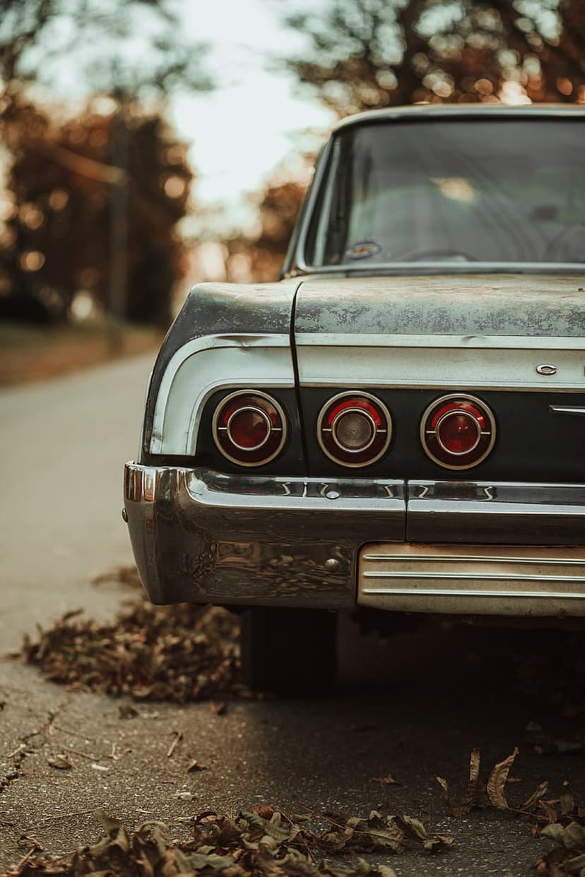 Triple red tail lights of a classic car above a chrome bumper on an asphalt road with dry autumn leaves.