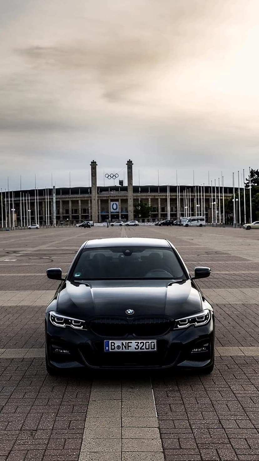Black BMW M3 parked on a stone plaza in front of the Berlin Olympic Stadium columns and iconic rings under gray clouds.