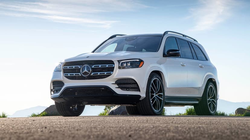 White Mercedes-Benz GLS luxury SUV parked on a mountain road with a clear blue sky and distant hills background.