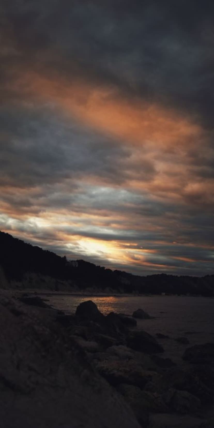 Rugged dark cliffs and a rocky shoreline under a cloudy twilight sky with orange reflections on the ocean water.