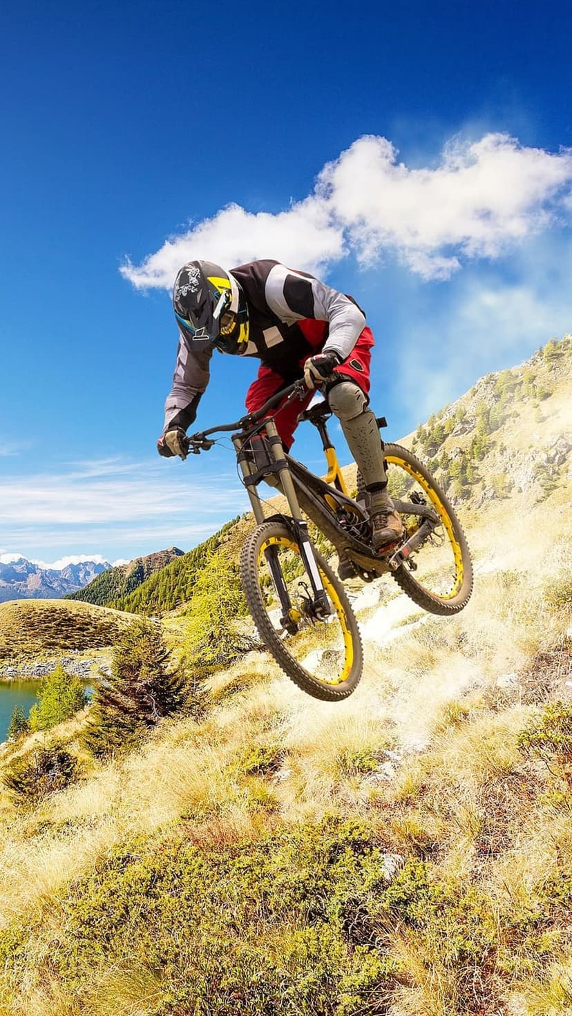 Mountain biker mid-jump over a steep rocky ridge with a turquoise alpine lake and jagged peaks in the distance.