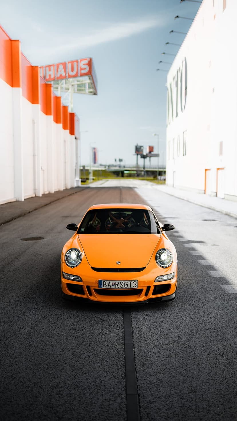 Orange Porsche 911 GT3 parked on an urban road between white and orange modern buildings under a clear blue sky.