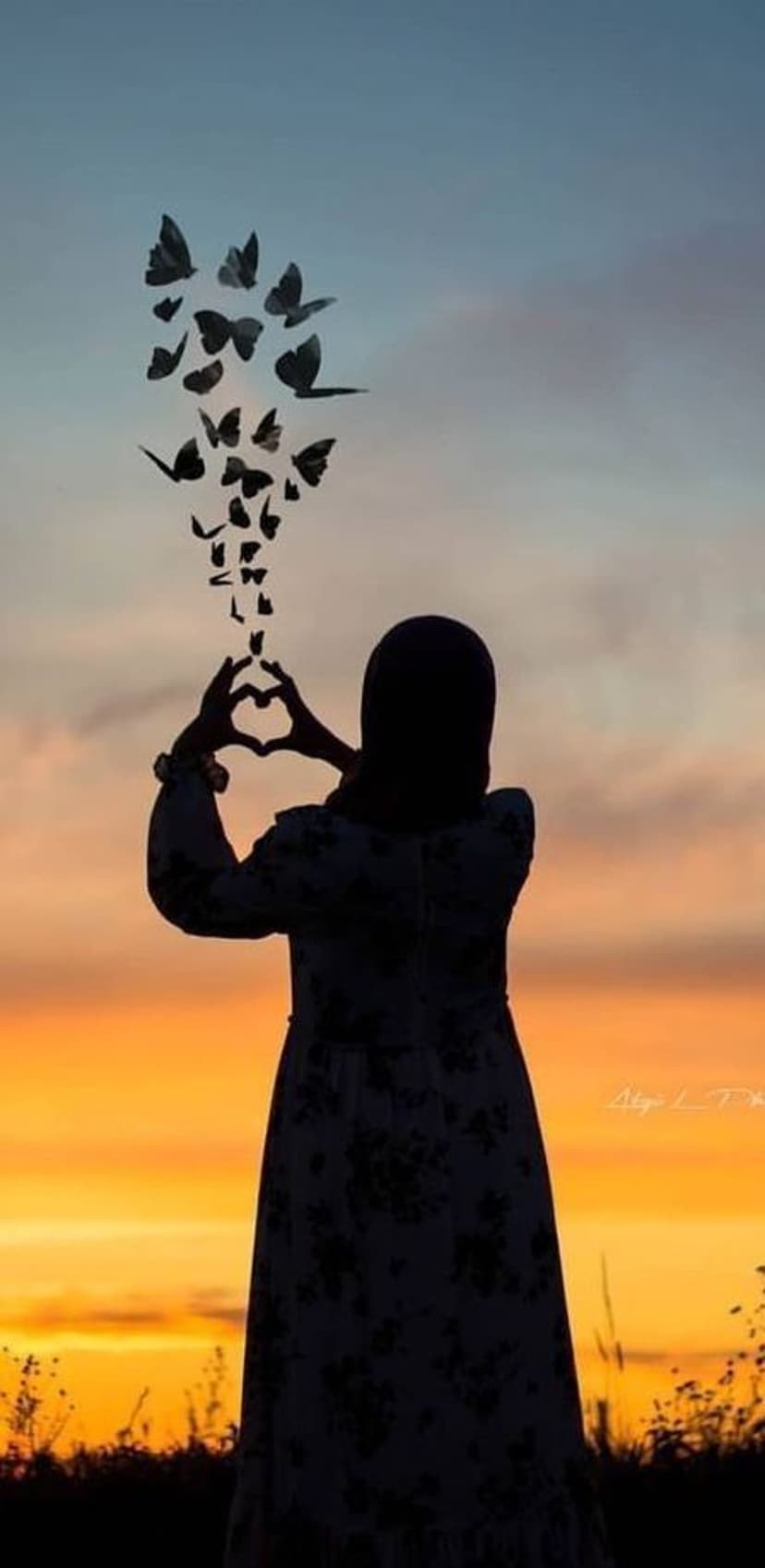 Woman in a hijab forms a heart with her hands as butterflies fly toward a vibrant orange and blue sunset sky.