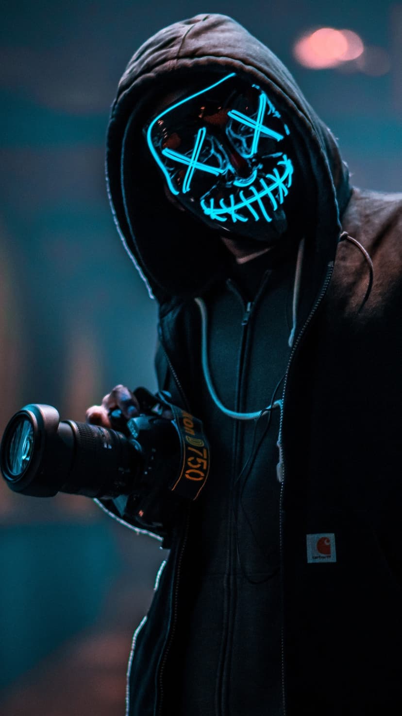 Masked photographer wearing a glowing blue neon mask with X eyes and holding a DSLR camera in a dark setting.