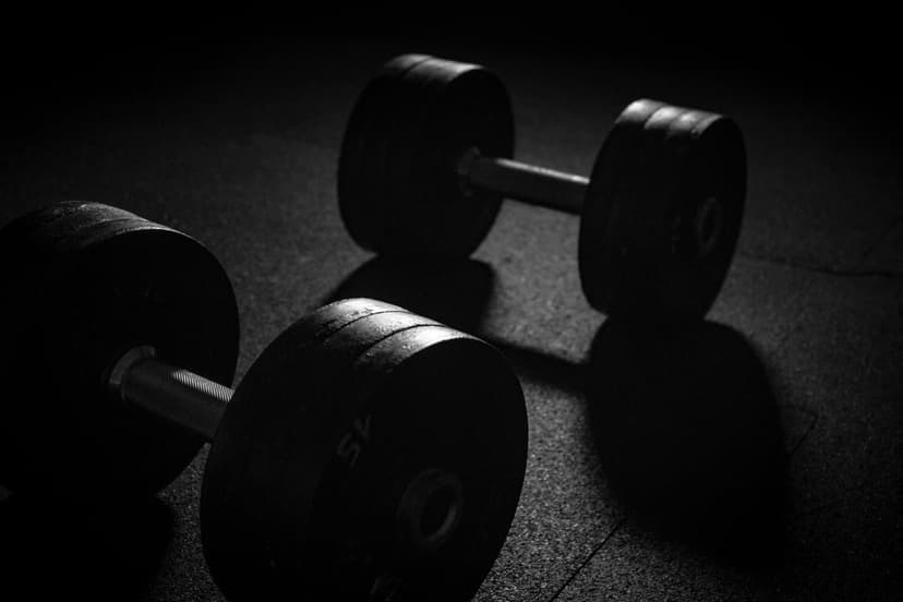 Two heavy dumbbells sit on a textured gym floor under harsh overhead lighting with deep black shadows.