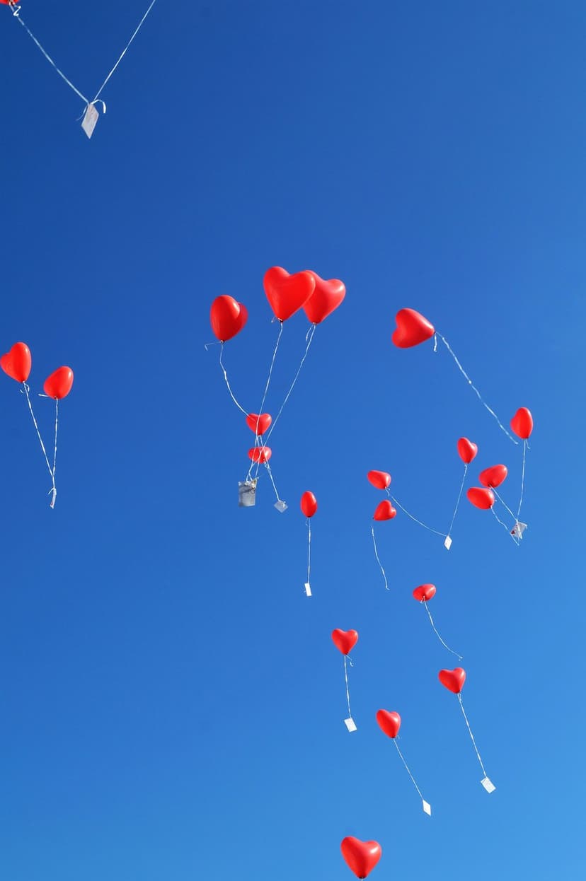 Red heart-shaped balloons with white strings ascend through a cloudless bright blue sky in bright daylight.