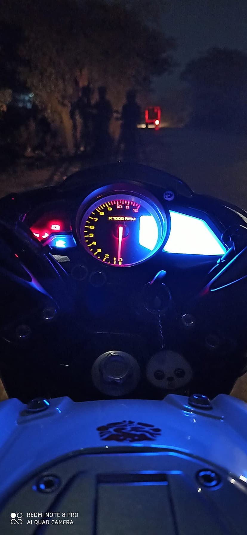Luminous blue motorcycle tachometer and dashboard glowing in the dark with red bokeh lights in the background.