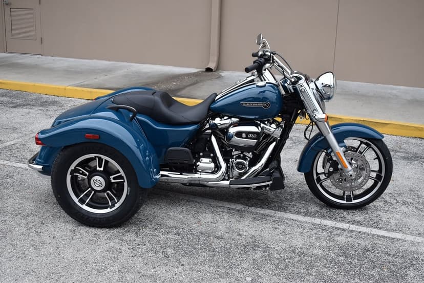 Blue Harley-Davidson trike with chrome engine and black leather seat parked against a beige wall on asphalt.