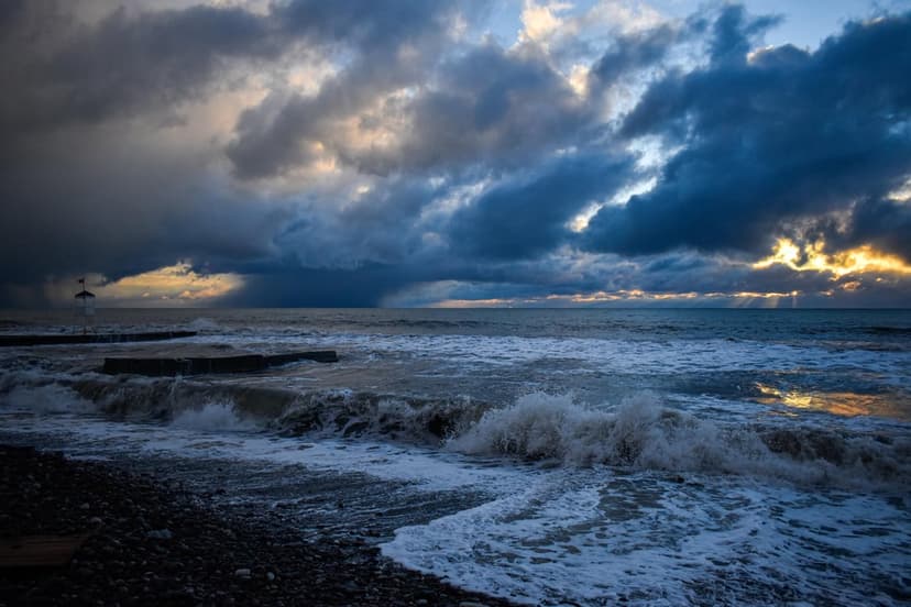Crashing white sea foam hits dark jagged rocks under a dramatic sunset sky with rays of light through clouds.