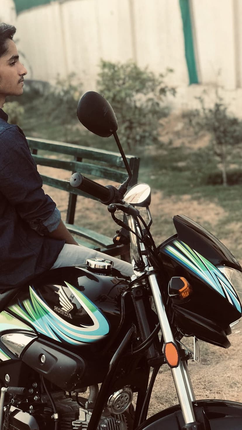 Young man in a denim shirt sits on a black Honda motorcycle with green graphics in a sunlit outdoor park.