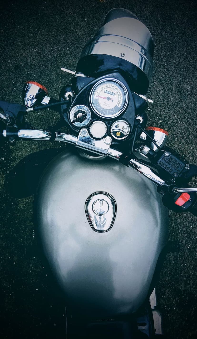 Classic silver motorcycle dashboard with chrome handlebars and a round speedometer viewed from above on asphalt.