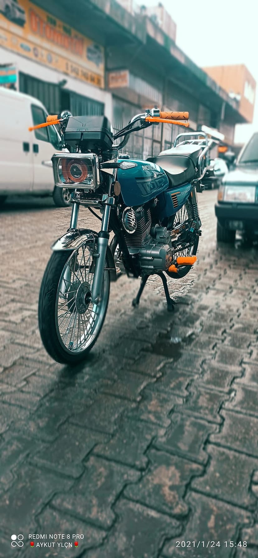 Teal motorcycle with orange accents parked on a wet, reflective city street under a gloomy, overcast sky.