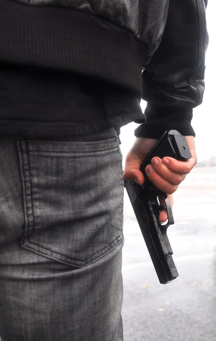 Man in a black bomber jacket and grey jeans holds a black pistol downward against his hip in an outdoor setting.