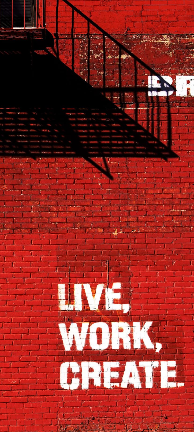 Red brick wall featuring white Live Work Create graffiti with dark shadows from a metal fire escape structure.
