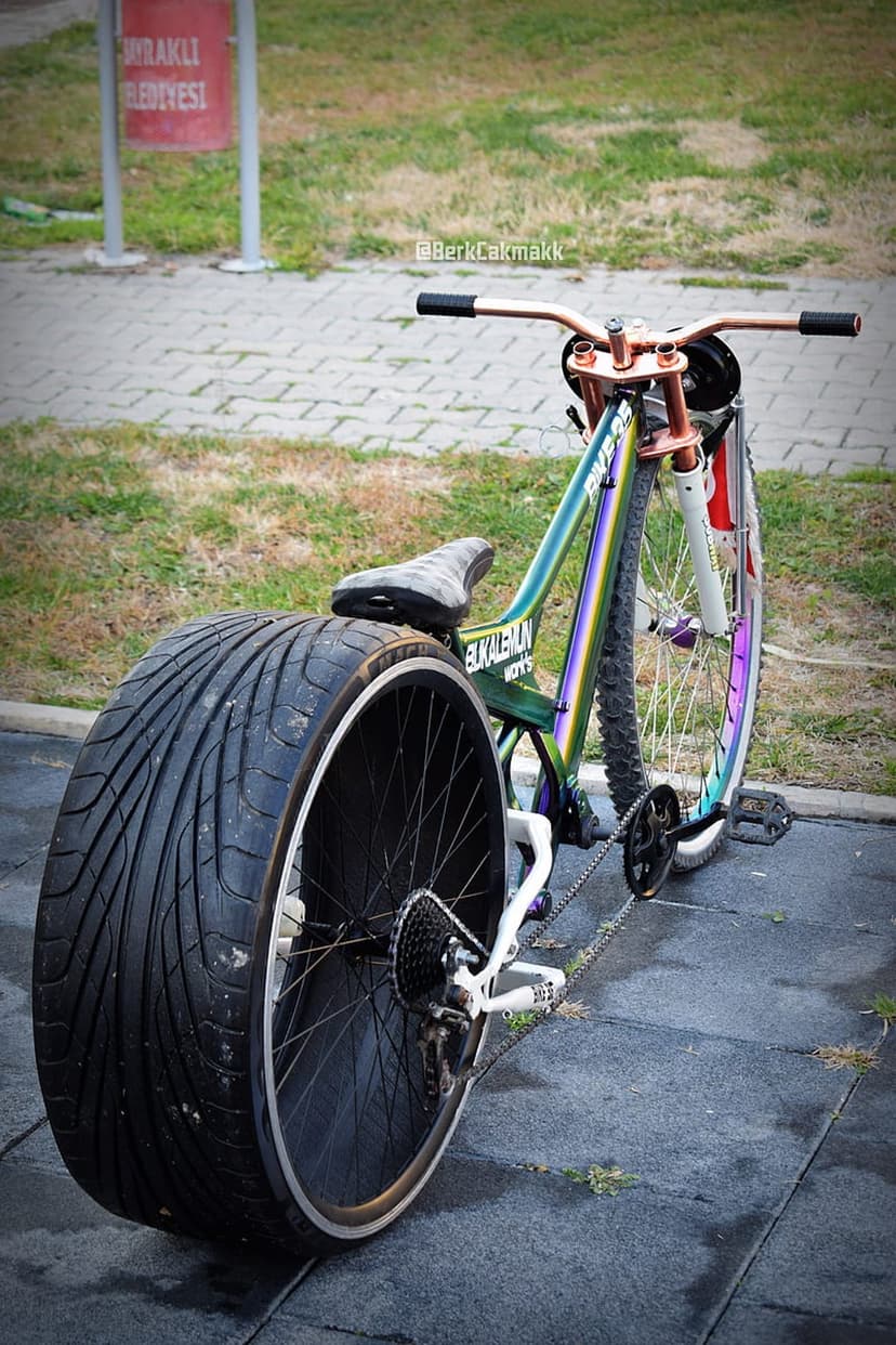 Custom iridescent bicycle with a huge car tire rear wheel and copper handlebars on a paved urban path.