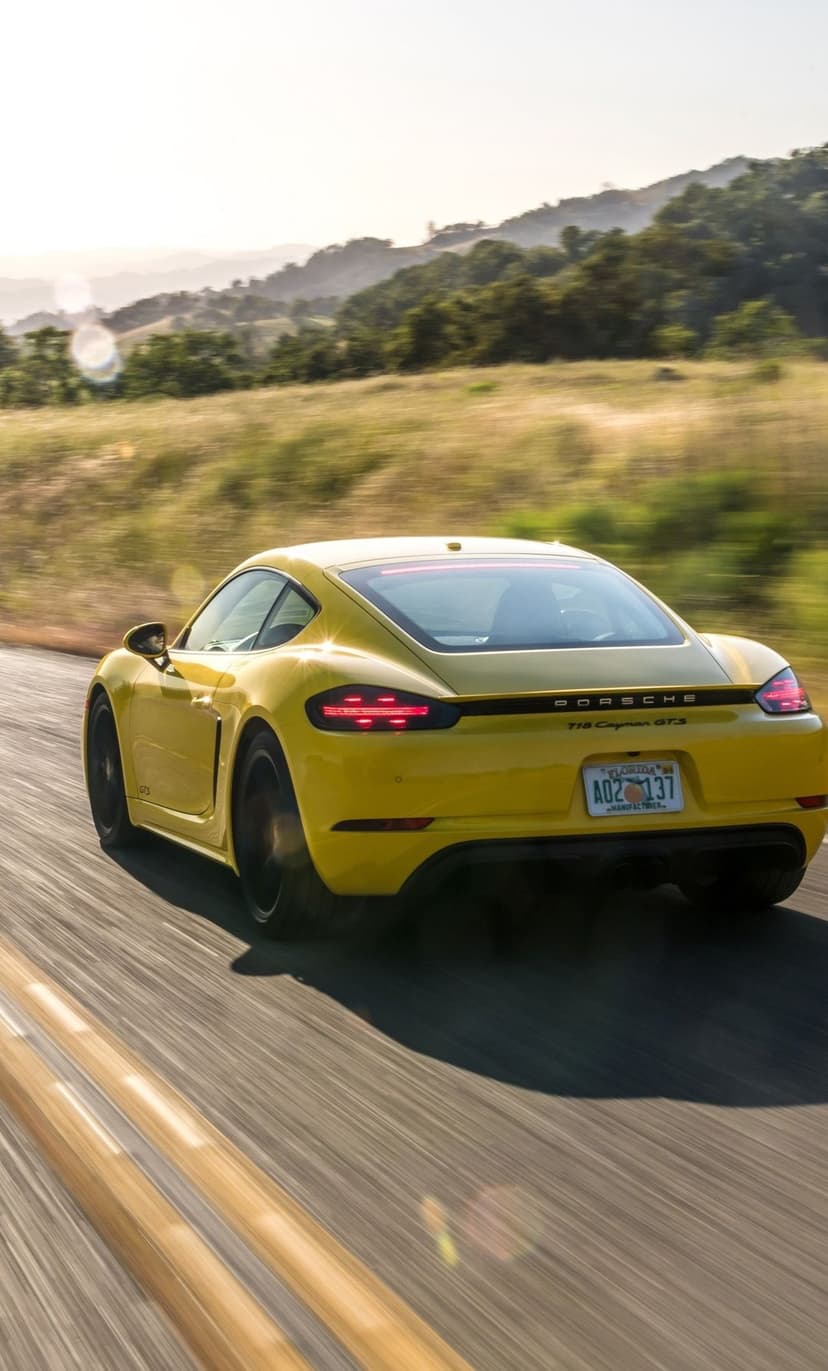 Yellow Porsche 718 Cayman GTS speeds along a road with motion blur against golden hills at sunset.