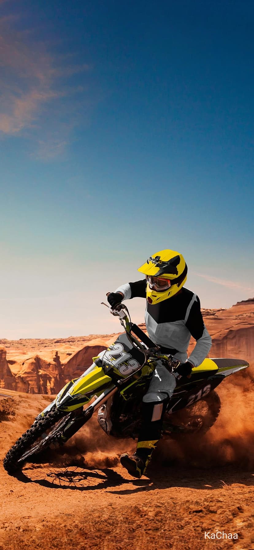 Motocross rider on a yellow bike leans into a dirt turn while kicking up dust in a rugged desert canyon.