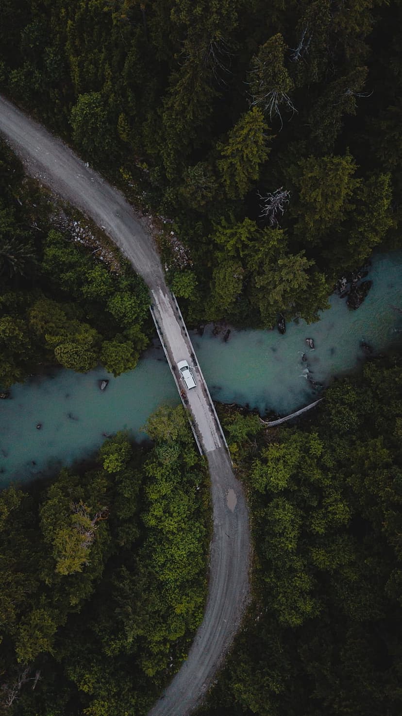 White car crossing a bridge over a bright turquoise river surrounded by dense dark green forest trees.