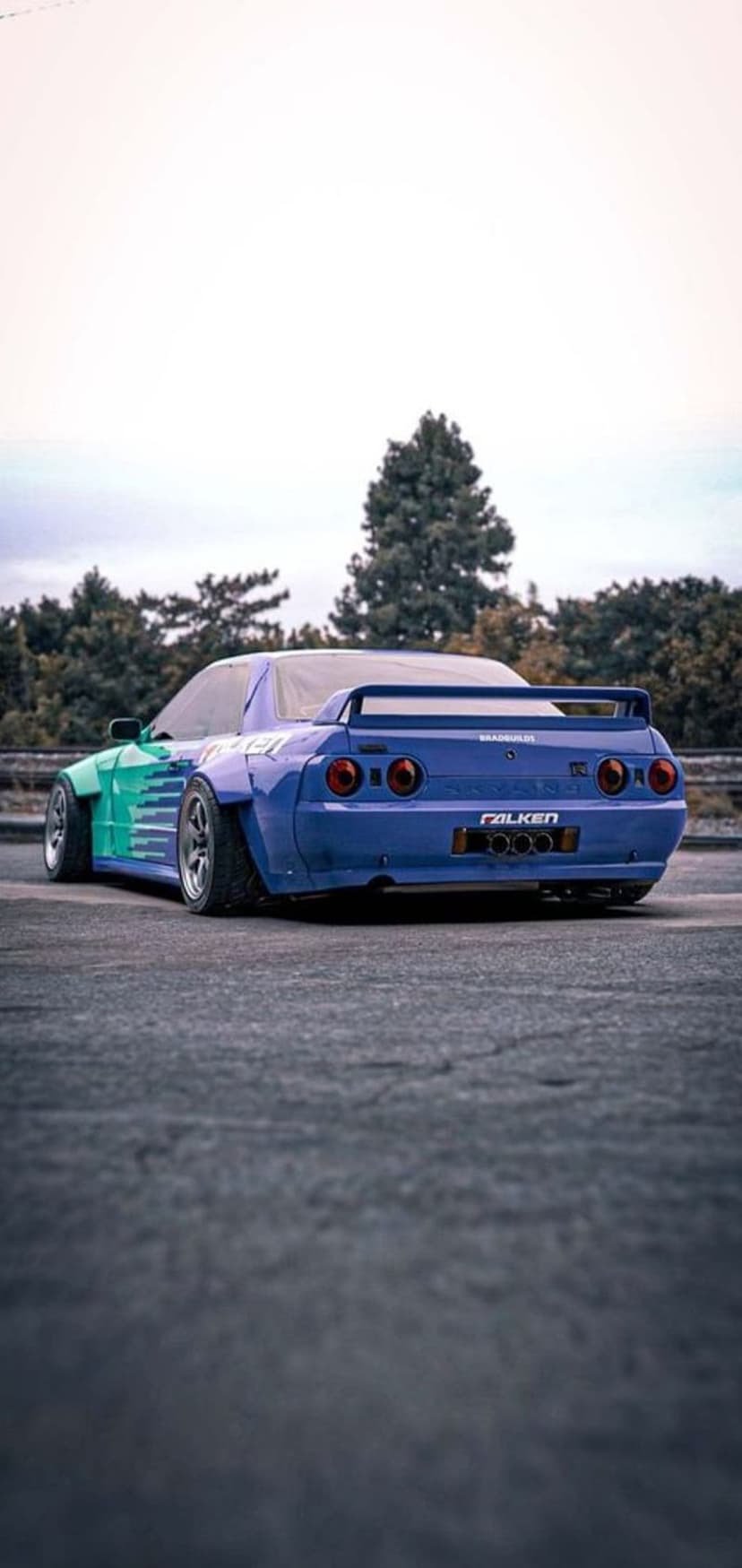 Nissan Skyline R32 with green and blue widebody kit and rear spoiler parked in front of overcast trees.