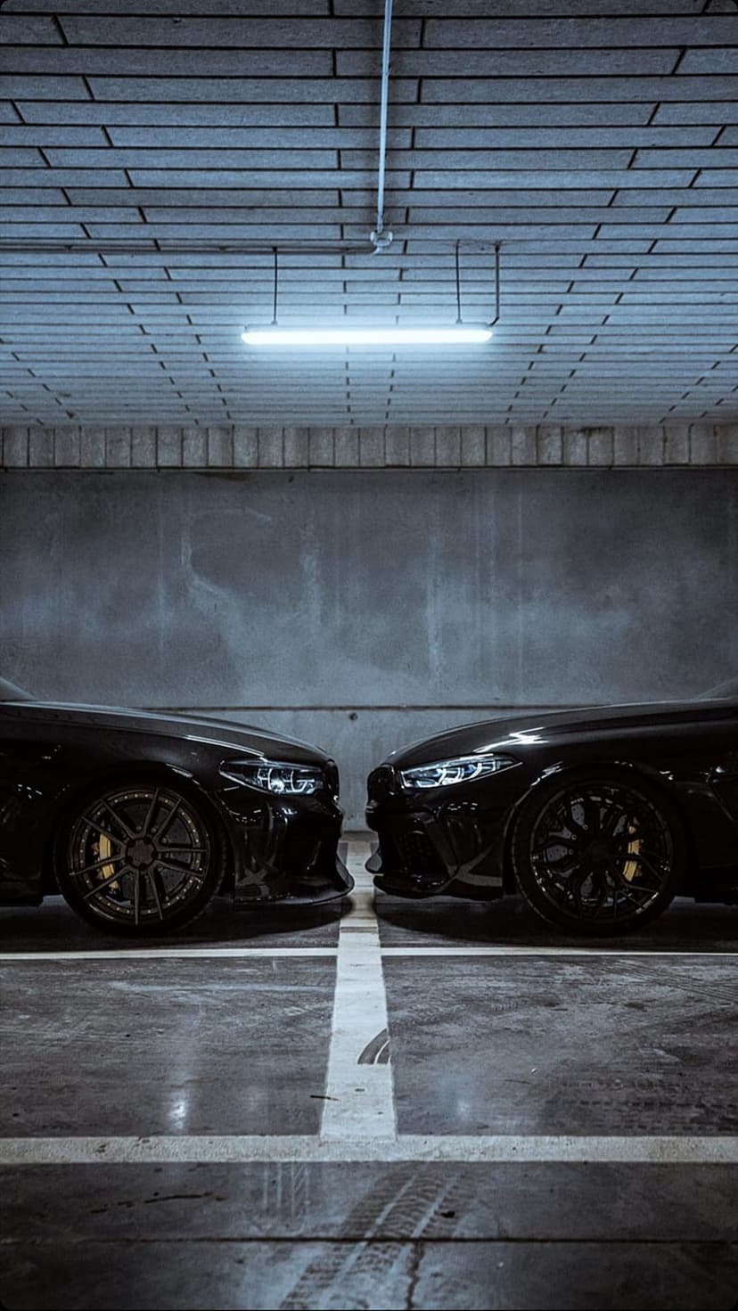 Two black luxury sports cars parked head-to-head in a dark garage with a blue fluorescent overhead light glow.
