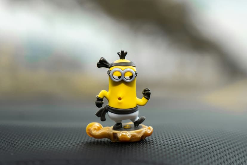 Yellow Minion toy in black ninja clothes stands on a sculpted golden cloud against a blurred background.
