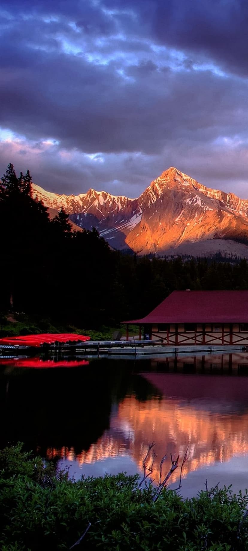 Snow-capped mountain peaks glow orange above a mirror lake with a red-roofed boathouse and parked red canoes.