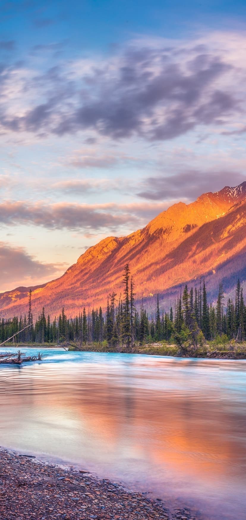 Fiery orange mountain peaks tower over a dark pine forest and a still river reflecting purple and blue sky tones.