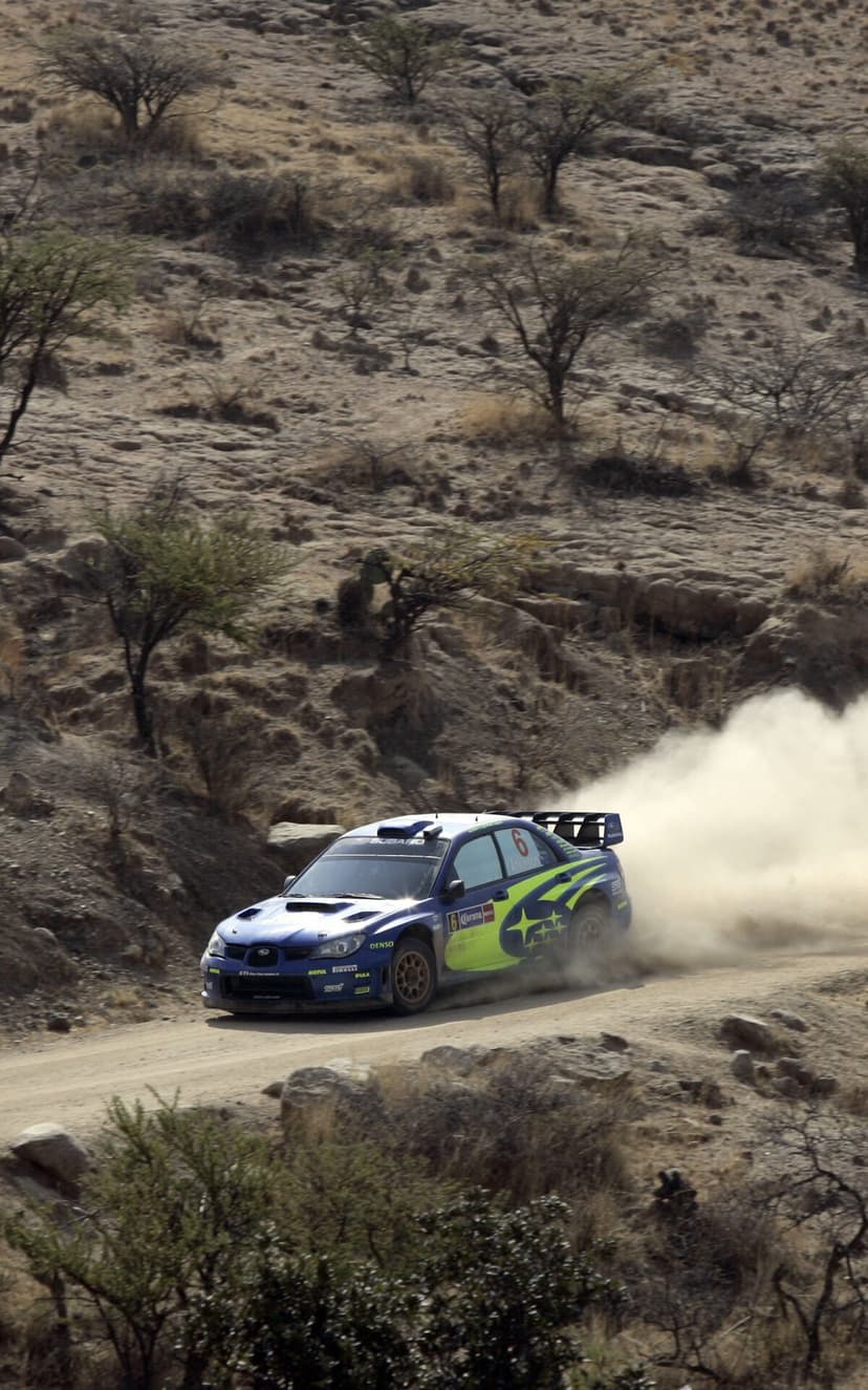 Blue Subaru Impreza WRC rally car kicks up a massive dust cloud on a dry, hilly dirt track in daylight.