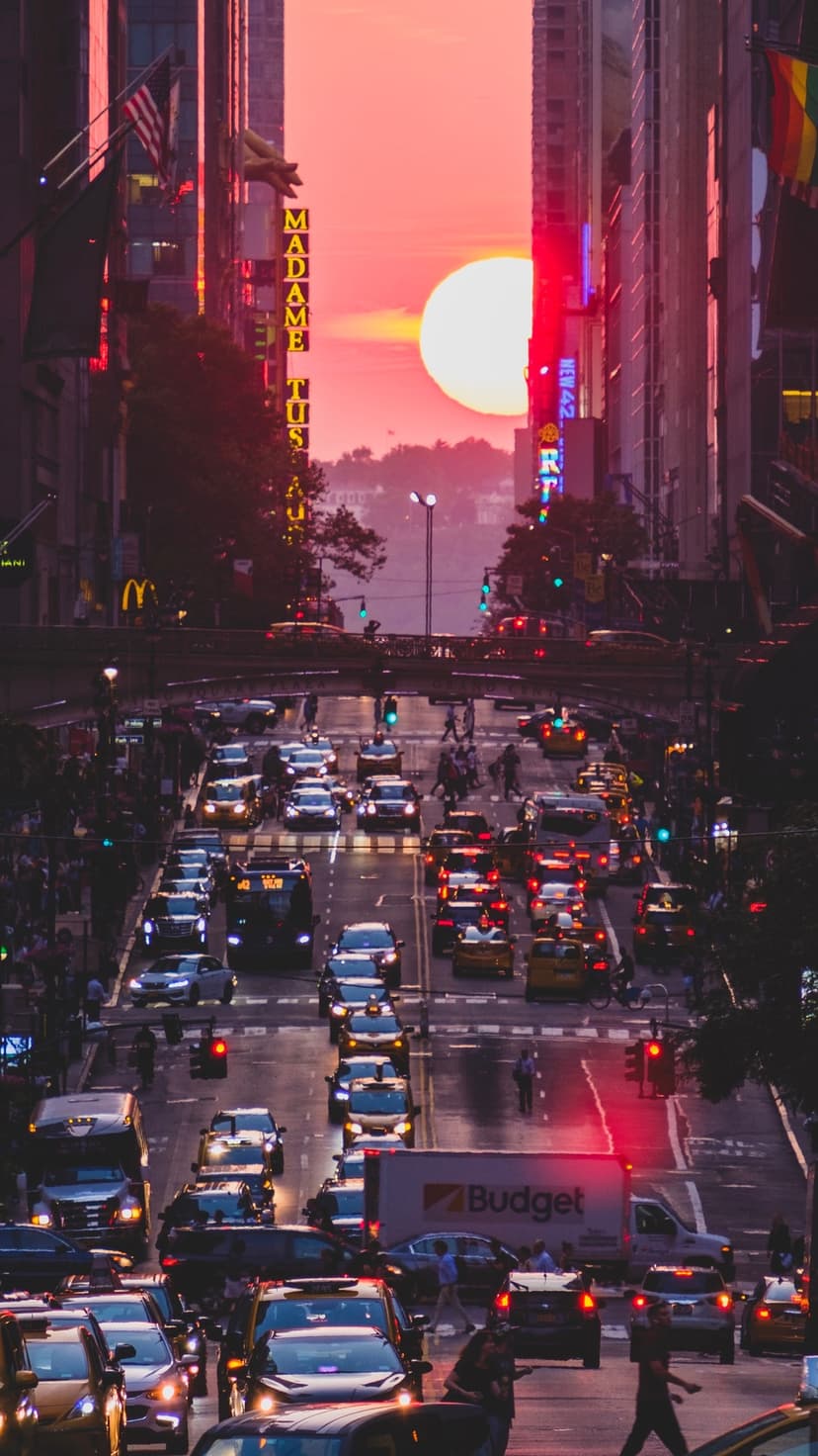 Fifth Avenue street traffic at sunset with a glowing sun between skyscrapers and bright neon city signs.