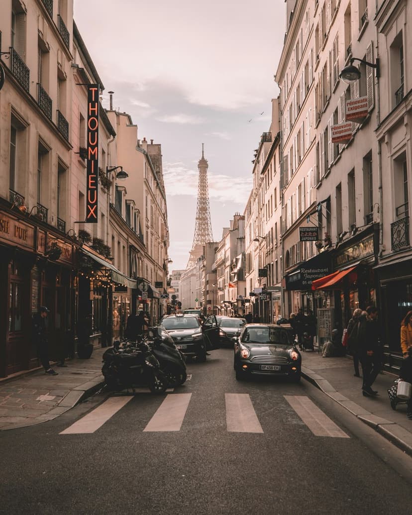 Eiffel Tower rises above a Parisian street lined with Haussmann buildings, parked cars, and shop storefronts.