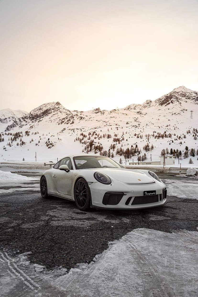 White Porsche 911 GT3 parked on a wet mountain road with snowy peaks and pine trees under a cloudy winter sky.