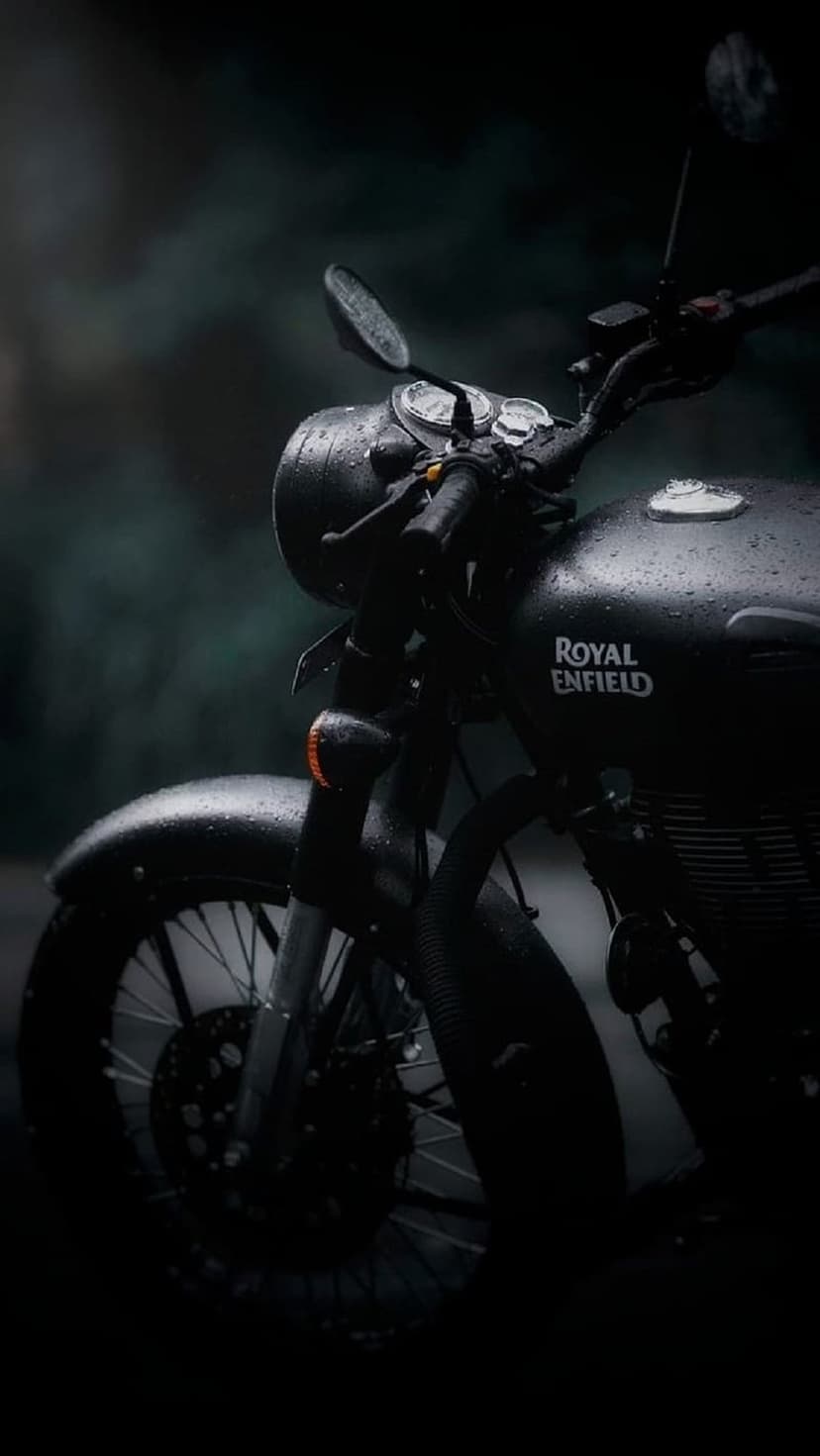 Black Royal Enfield motorcycle with water droplets on the fuel tank and headlight during a rainy night outdoors.