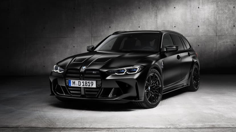 Front view of a matte black BMW M3 Touring wagon in a dark concrete studio with glowing LED headlights.