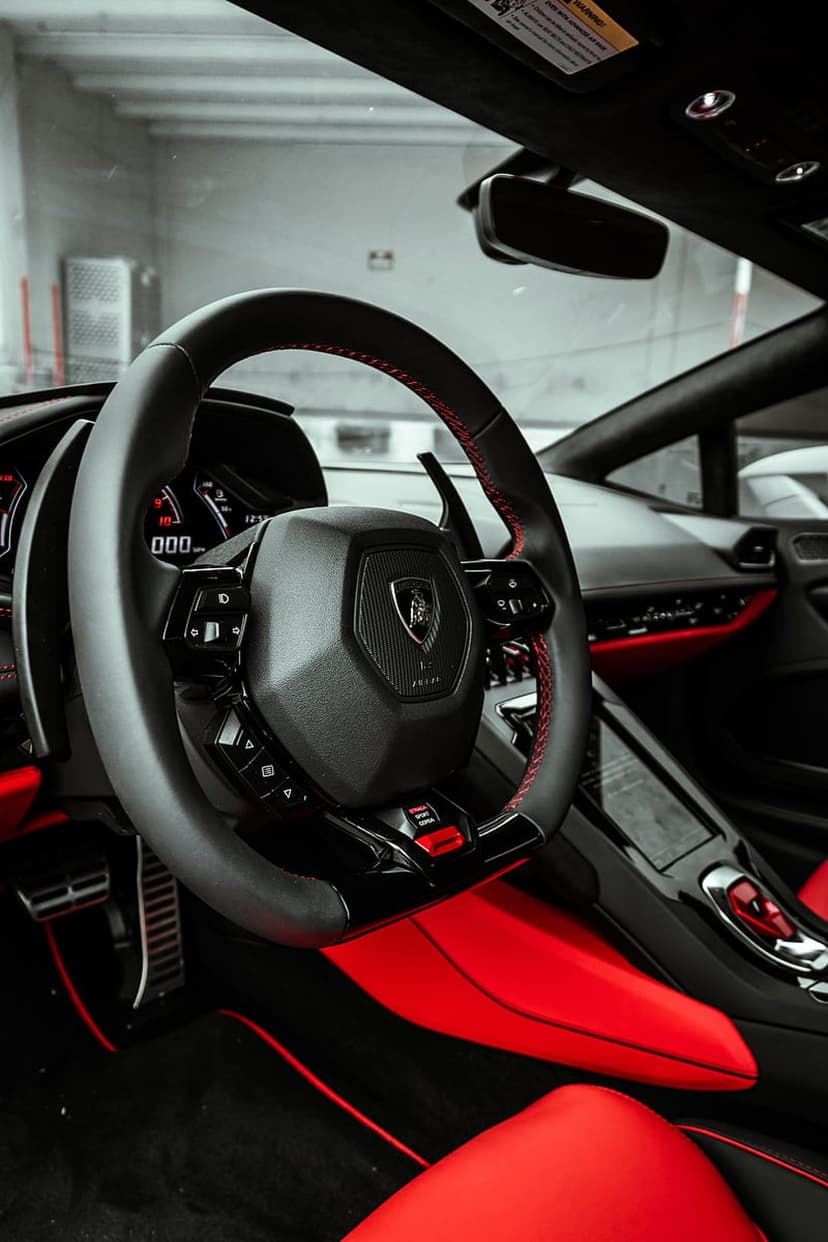 Lamborghini steering wheel with red stitching and emblem inside a black and crimson luxury supercar cockpit.