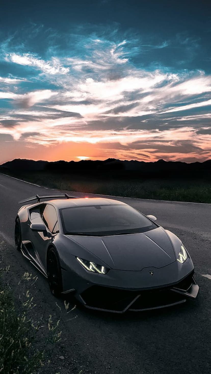Dark gray Lamborghini supercar parked on a roadside with orange and purple sunset clouds over mountain silhouettes.