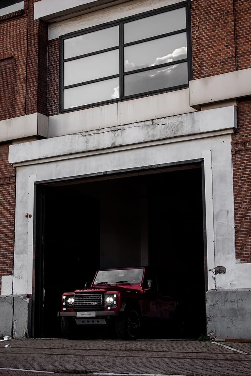 Red Land Rover Defender partially inside a dark brick garage under a multi-paned window reflecting a cloudy sky.