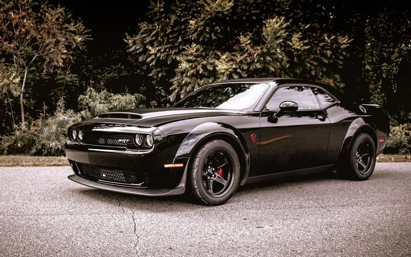 Black Dodge Challenger SRT Demon parked on a cracked asphalt road against a background of dark green trees.