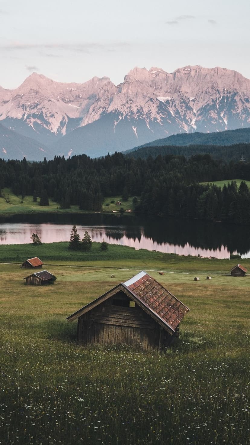 Weathered wooden barns sit in a lush green meadow before a reflective lake and towering snow-dusted mountains.