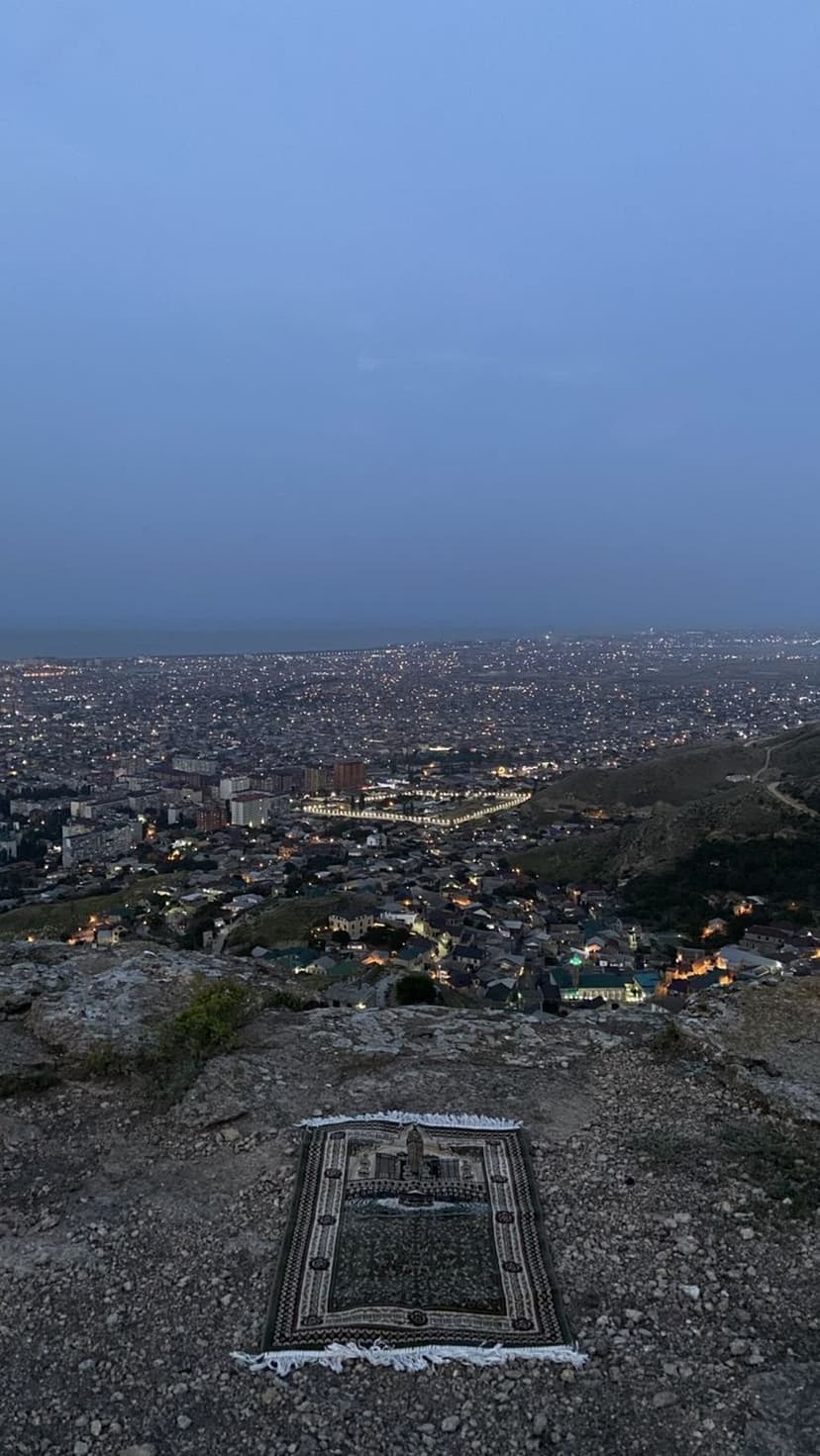 Prayer mat on a rocky cliff overlooking a vast city glowing with golden lights under a blue twilight sky.