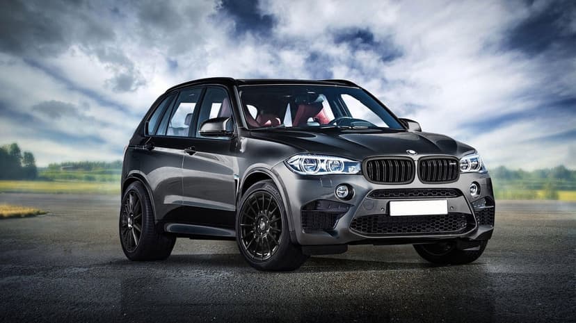 Gray BMW X5 M performance SUV with red seats and black wheels parked on a wet road under cloudy skies.