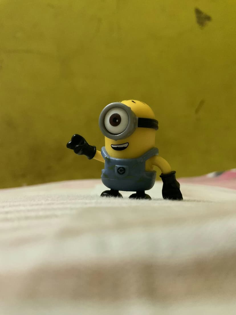 One-eyed Minion toy wearing blue overalls waves a hand while standing on a white and yellow textured background.