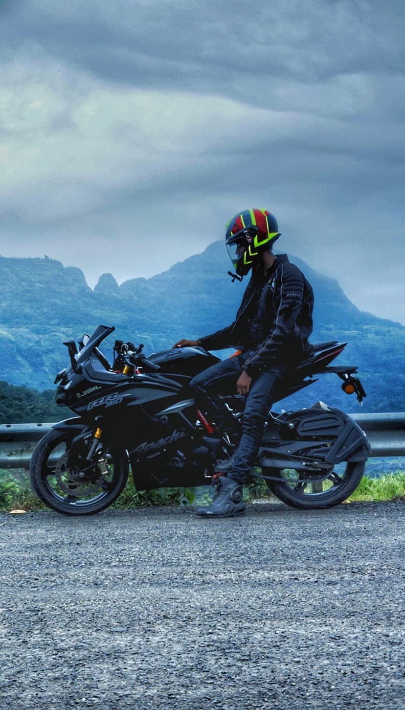 Motorcyclist in a bright helmet sits on a black bike with cloudy mountain peaks visible in the background.