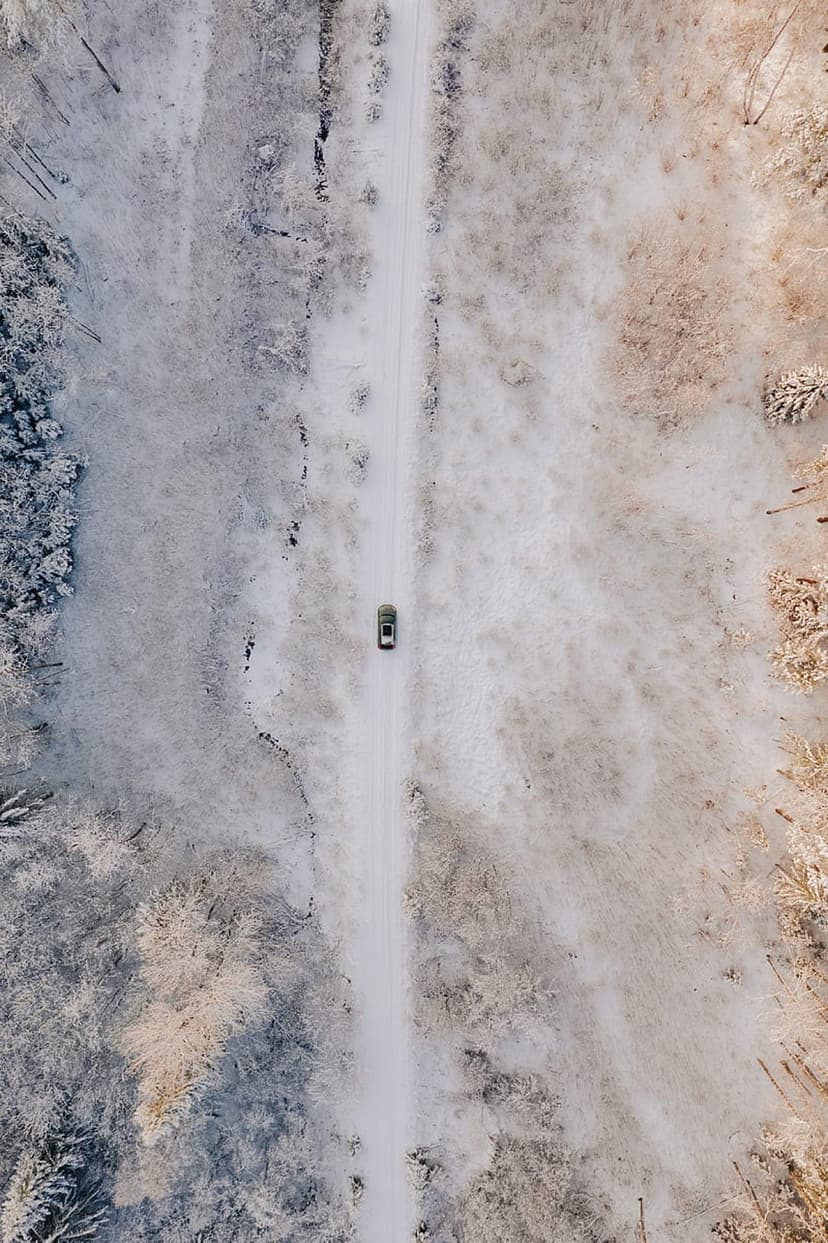 Dark car drives along a narrow snowy road through a frosted pine forest captured from a top-down drone angle.